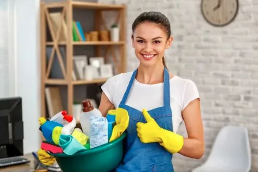 A woman stands holding a bucket filled with various cleaning supplies, ready to tackle her cleaning tasks.