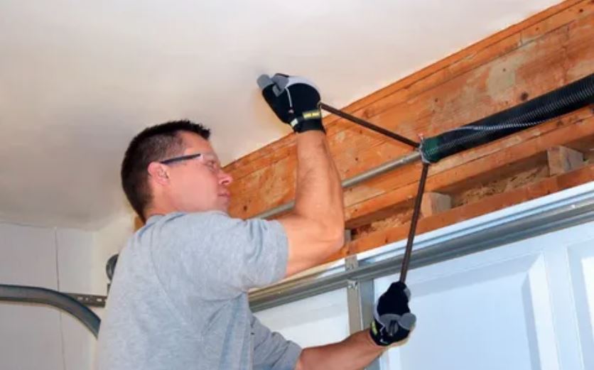 A man is repairing a garage door, focused on his task with tools in hand, surrounded by a garage setting.